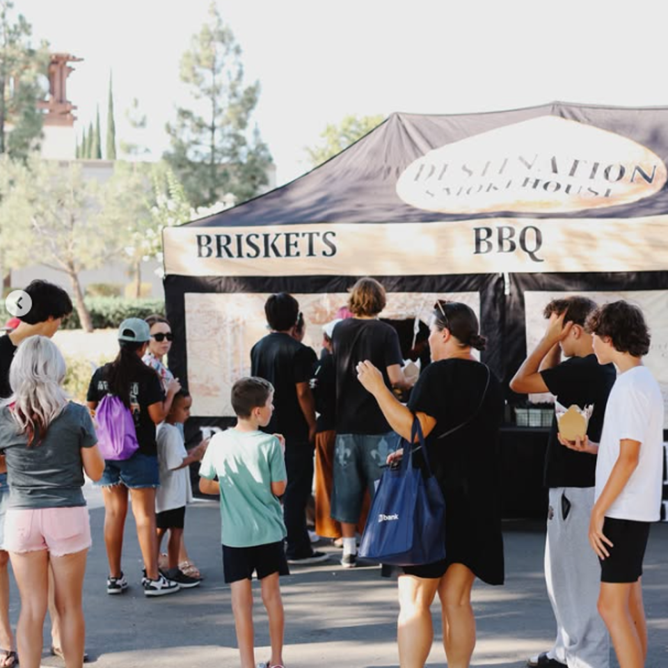 Crowd gathers at an outdoor barbecue stand advertising briskets and BBQ, with people ordering beneath a branded canopy.