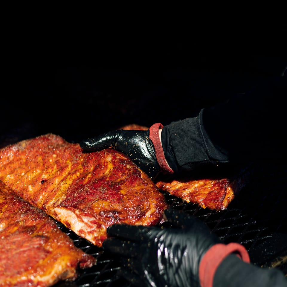 Gloved hands arrange seasoned barbecue ribs on a hot grill.