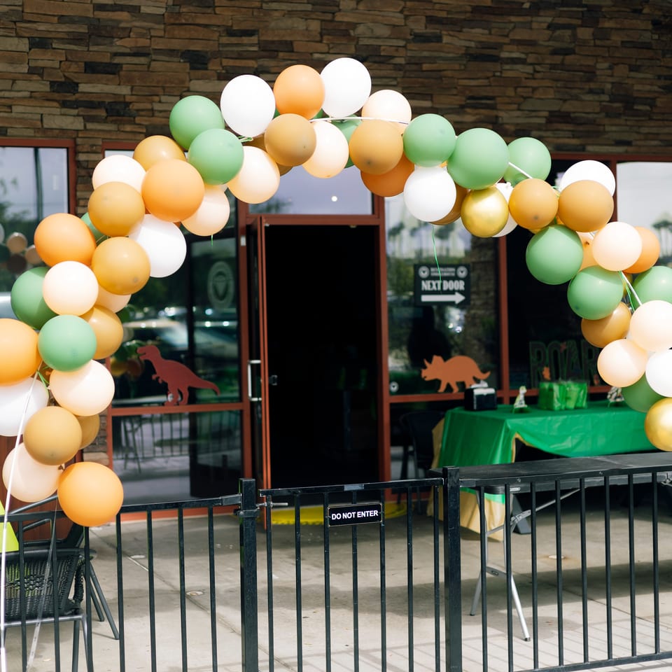 Restaurant entrance decorated with a colorful balloon arch, green table, and dinosaur silhouettes behind a metal barrier.