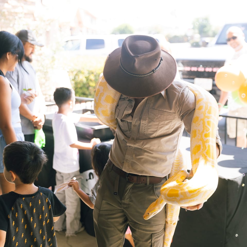Entertainer wearing a cowboy hat holds a large yellow-and-white snake while children and adults watch at an outdoor event.