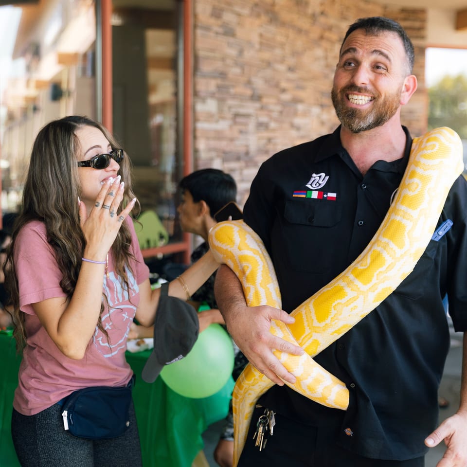 Smiling man in black attire holds a large yellow-and-white patterned snake at a lively indoor gathering.