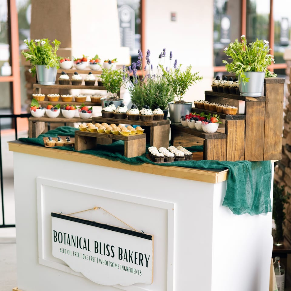 Botanical Bliss Bakery display table featuring cupcakes, pastries, strawberries, potted herbs, and flowers in a bright café setting.