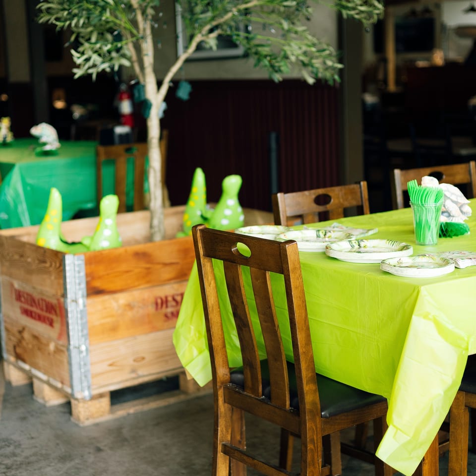 Restaurant dining area decorated with a lime-green tablecloth, wooden chairs, leafy tree, plates, and colorful ceramic ornaments.